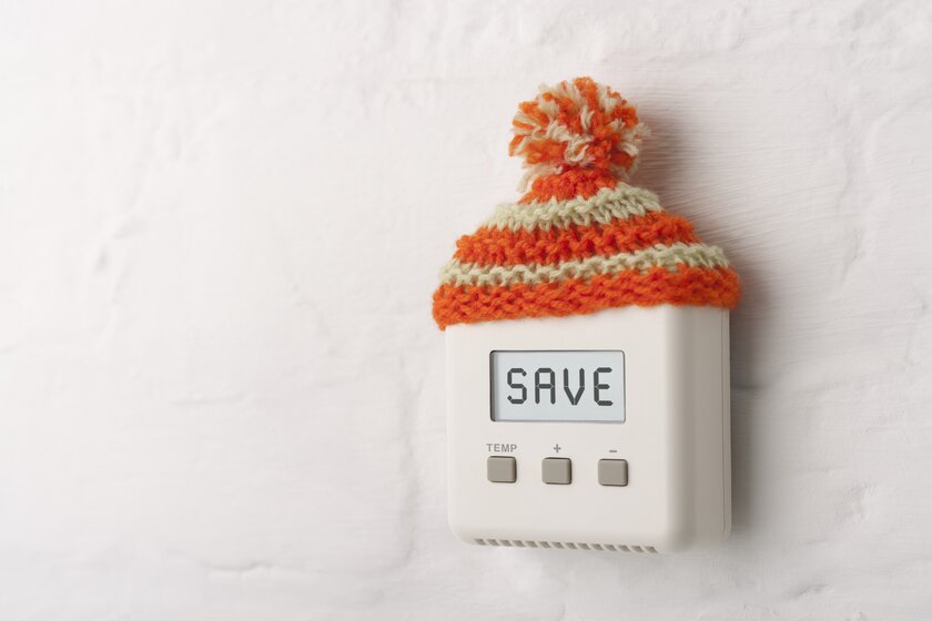 Thermostat trägt eine Wollmütze und enthält als Anzeige die wörtliche Aufforderung zum Sparen!