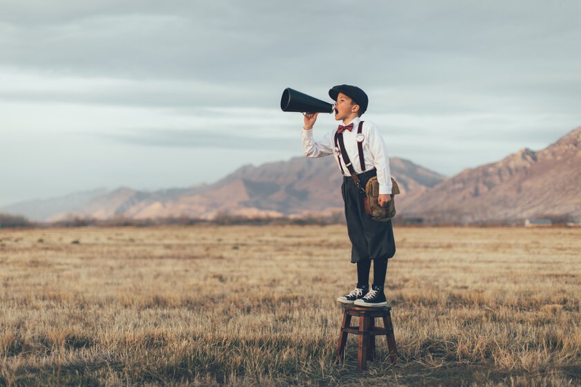 A news boy dressed in vintage knickers and newsboy hat stands yelling through a megaphone in the middle of a field in Utah, USA. 