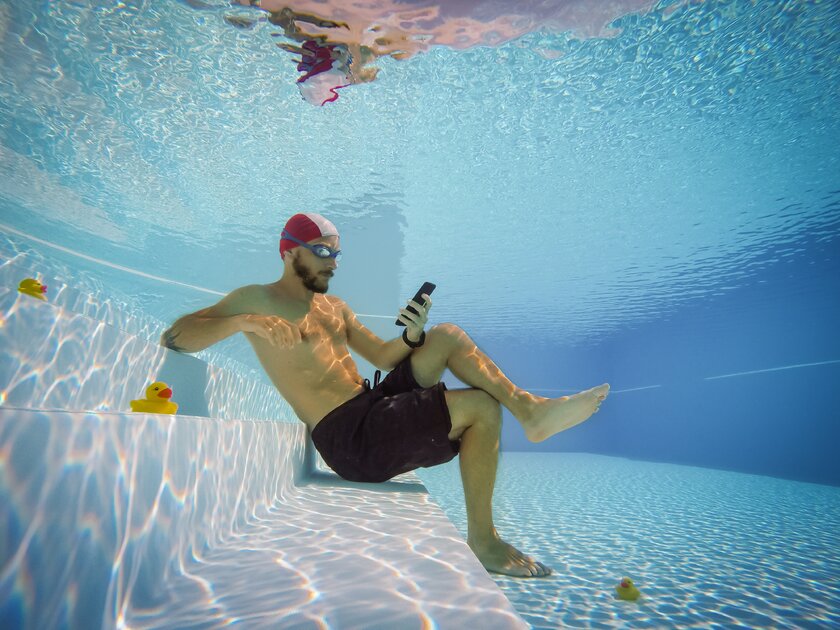Mann in Badehose und Schwimmbrille sitzt entspannt unter Wasser in einem Pool und schaut gebannt auf sein Smartphone.