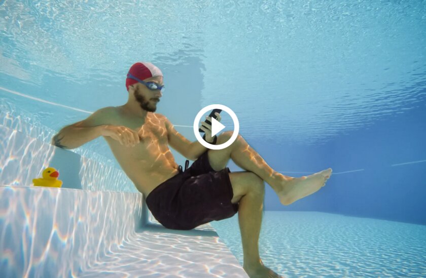 Mann in Sportbadekleidung sitzt unter Wasser und schaut gebannt auf sein Smartphone.
