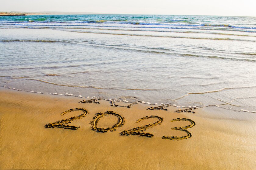 Meereswellen überspülen in Sand geschriebene Jahreszahlen für 2022, dahinter sichtbar bleibend der Schriftzug für das neue Jahr 2023.