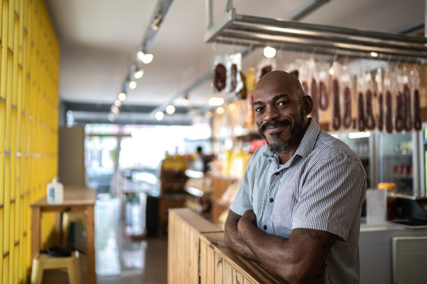 Männlicher Unternehmer lehnt lächelnd über seinen Verkaufstresen im Lebensmittelgeschäft.