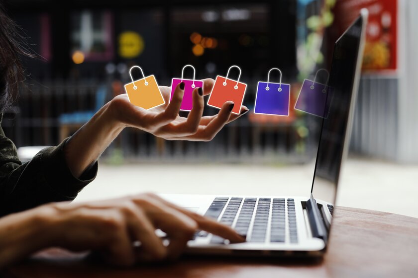 Weibliche Hand an einem Laptop entlässt virtuelle Einkaufstüten in vor ihr befindlichen Bildschirm.