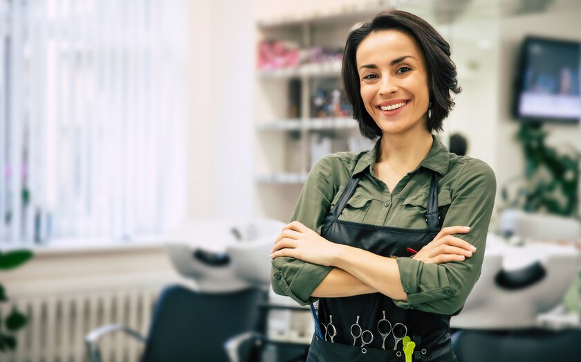 Smiling hairdresser stands in her salon and wears belt with many scissors.