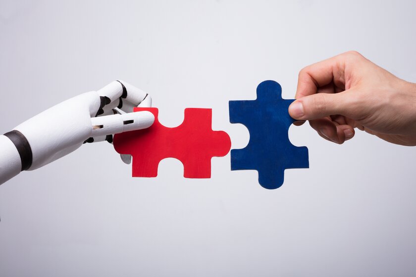 Roboterhand mit Puzzleteil von links fügt sich in Puzzleteil aus Menschenhand von rechts.