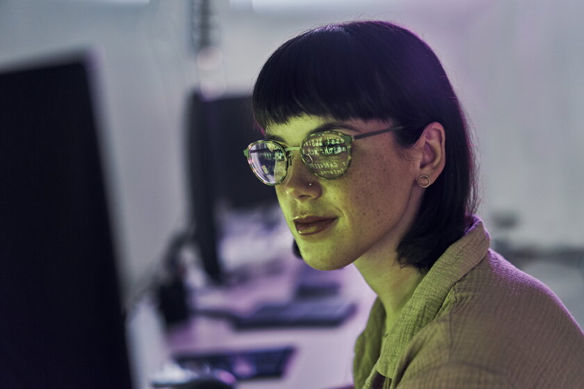Frau blickt auf Computerbildschirm mit Programmiercode in ihren Brillenglaesern widerspiegelnd.