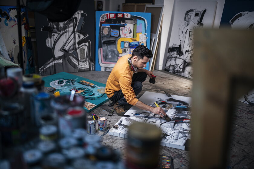 Maler in seinem Atelier umgeben von Bildern bei der Bearbeitung neuer Kunstwerke.