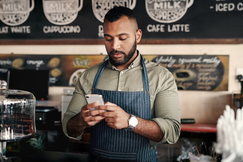 Besitzer eines Coffeeshops lehnt am Tresen und blickt auf das Smartphone in seiner Hand.