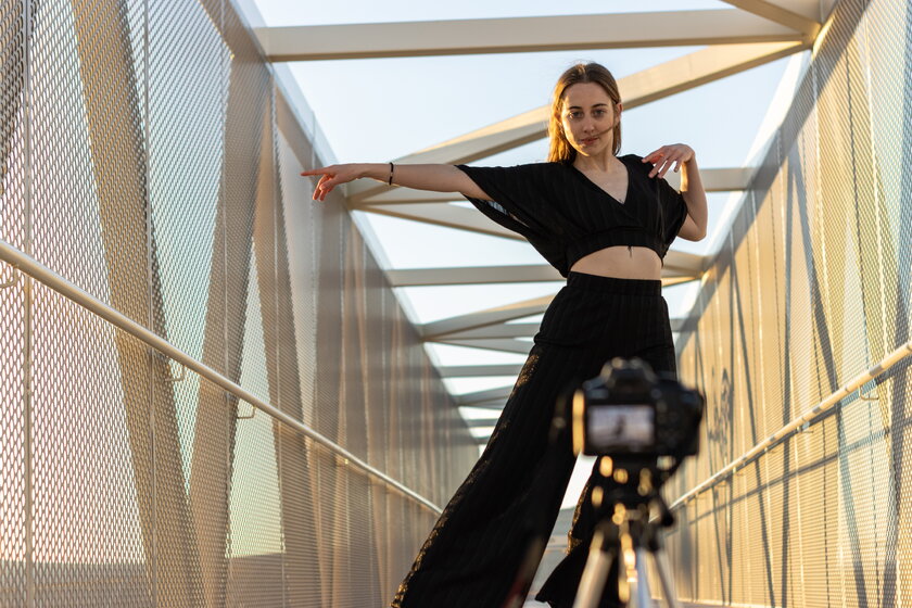 Frontaler Blick auf eine tanzende junge Frau vor einer Videokamera auf einer Bruecke.