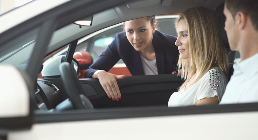 Nahaufnahme der Seitenansicht eines Paares mittlerer Erwachsener, das in einem neuen Auto sitzt, die Verkaeuferin lehnt sich durch das Beifahrerfenster.