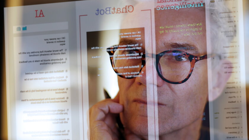 Gesicht eines Mannes mittleren Alters mit Brille und Stift haltend, gespiegelt auf Chatbot-Bildschirm.