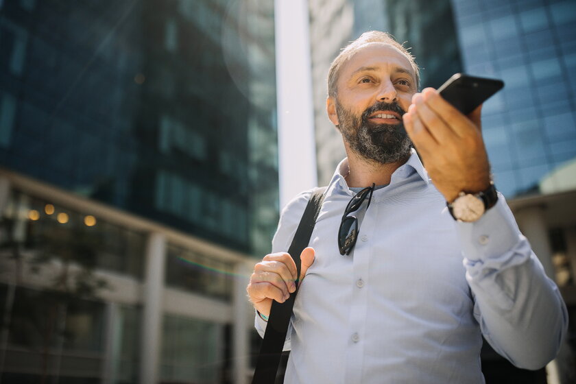 Mann am Smartphone vor Wolkenkratzer nutzt Sprachfunktion unterwegs.